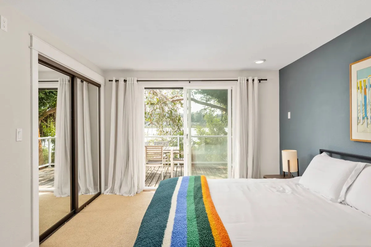 A serene bedroom with a colorful blanket on the bed, large glass doors opening to a quiet outdoor view, and a mirrored closet. The room is tranquil, filled with natural light, and offers a restful escape after a day of lakeside adventures with pets.