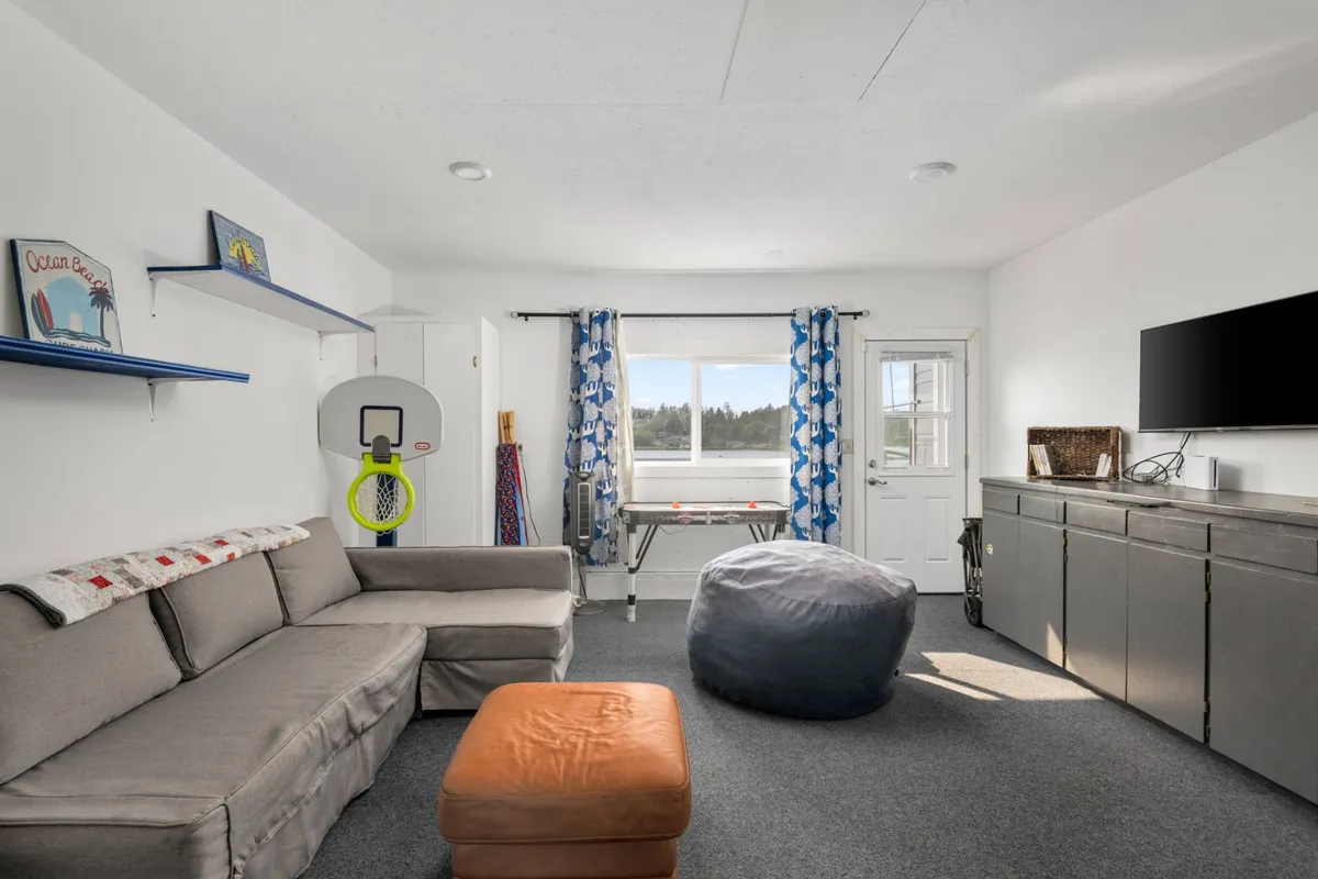 Inside, the image reveals a game room with a comfortable sofa, bean bag, a basketball hoop, TV, and window views. The layout suggests flexibility for various activities, from game nights to movie marathons and playful afternoons, perfectly fitting a multi-purpose space for all ages.