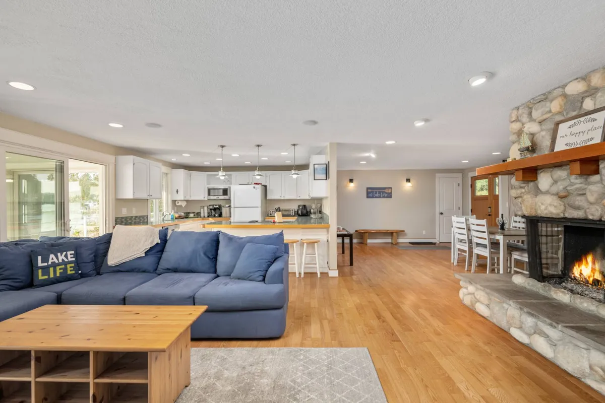 This image captures an open-plan interior with a large blue sectional sofa, a stone fireplace, a dining table, and a kitchen area. The space is arranged for fluid movement between living and dining areas, with cozy seating and a warm, welcoming atmosphere conducive to socializing and family meals.