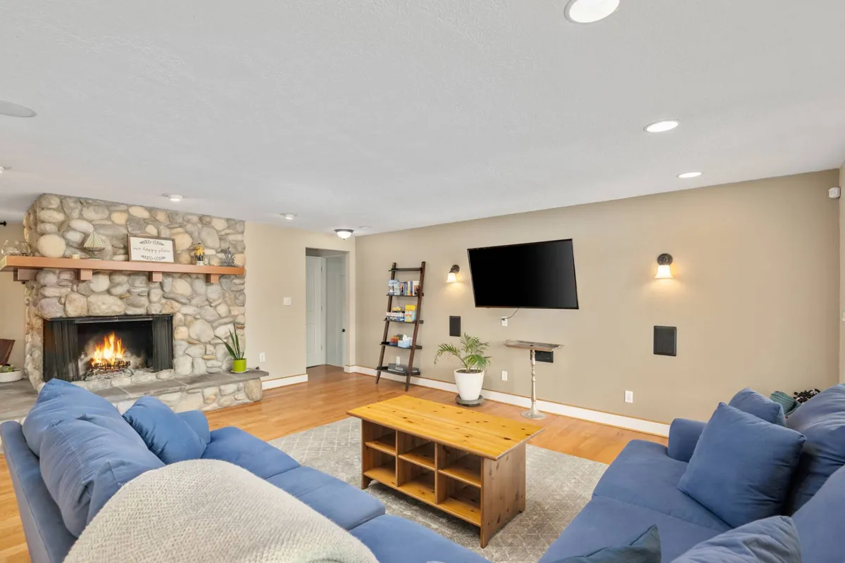 A spacious living area with blue sofas arranged around a stone fireplace, a wooden coffee table at the center, and a wall-mounted TV. The room is warmly lit and inviting, with ample floor space for pets to relax and families to gather for cozy evenings by the fire.