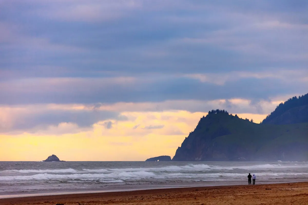 A breathtaking coastal scene with ocean waves rolling against the shoreline, distant cliffs, and a couple strolling on the beach. The mood is peaceful and romantic, evoking the spirit of the Oregon Coast’s natural wonders.