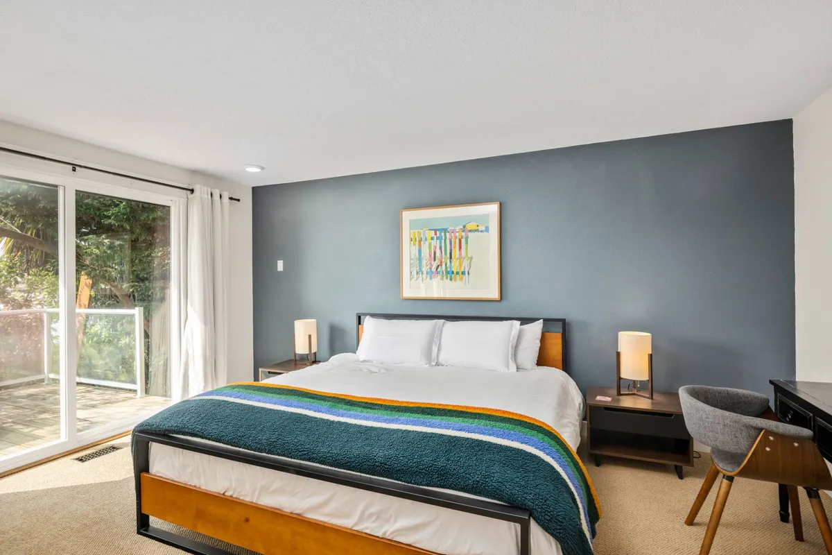 A serene bedroom featuring a plush bed with crisp linens, tasteful artwork above the headboard, nightstands, a comfy chair, and a sliding glass door that lets in natural light and provides a glimpse of the outdoors. The overall mood is tranquil, with soft colors and a sense of privacy and relaxation.