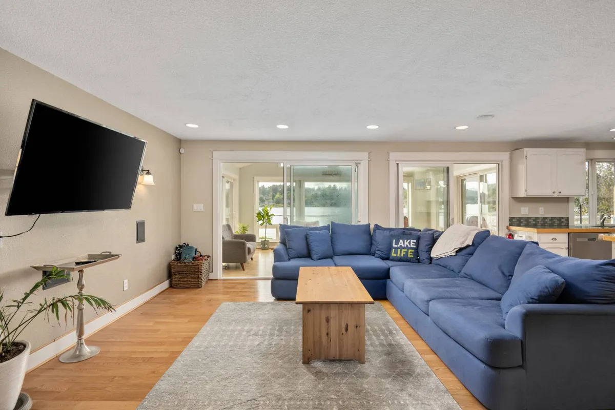 This image shows a spacious living room featuring a large blue sectional sofa, a wooden coffee table, a television, and expansive windows that deliver panoramic views of Devil's Lake. The room is bathed in natural light, highlighting the plush furnishings and the inviting atmosphere ideal for gatherings.