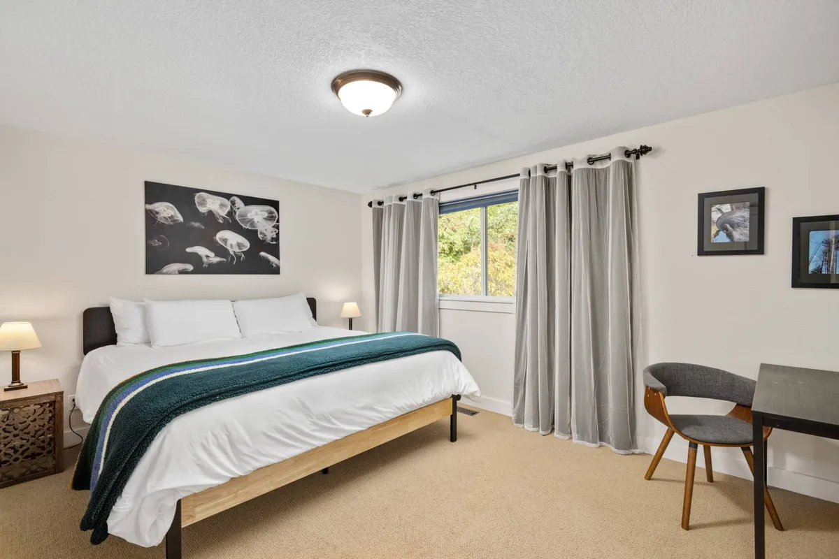 A peaceful bedroom with a bed dressed in white linens and a teal throw, a window flooding the room with daylight, jellyfish artwork above the bed, and a cozy reading nook. The space feels quiet and inviting, perfect for unwinding after a day of fun.