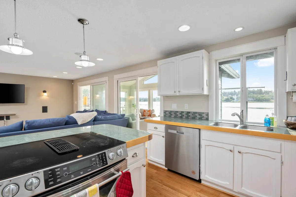 This image captures a vibrant kitchen scene centered on the kitchen island, with a blue couch and large windows framing expansive water views. The atmosphere is cheerful and relaxed, with hints of cooking and entertaining activity. The open layout connects the kitchen visually with the living space and outdoors, emphasizing both culinary action and lakeside ambiance.