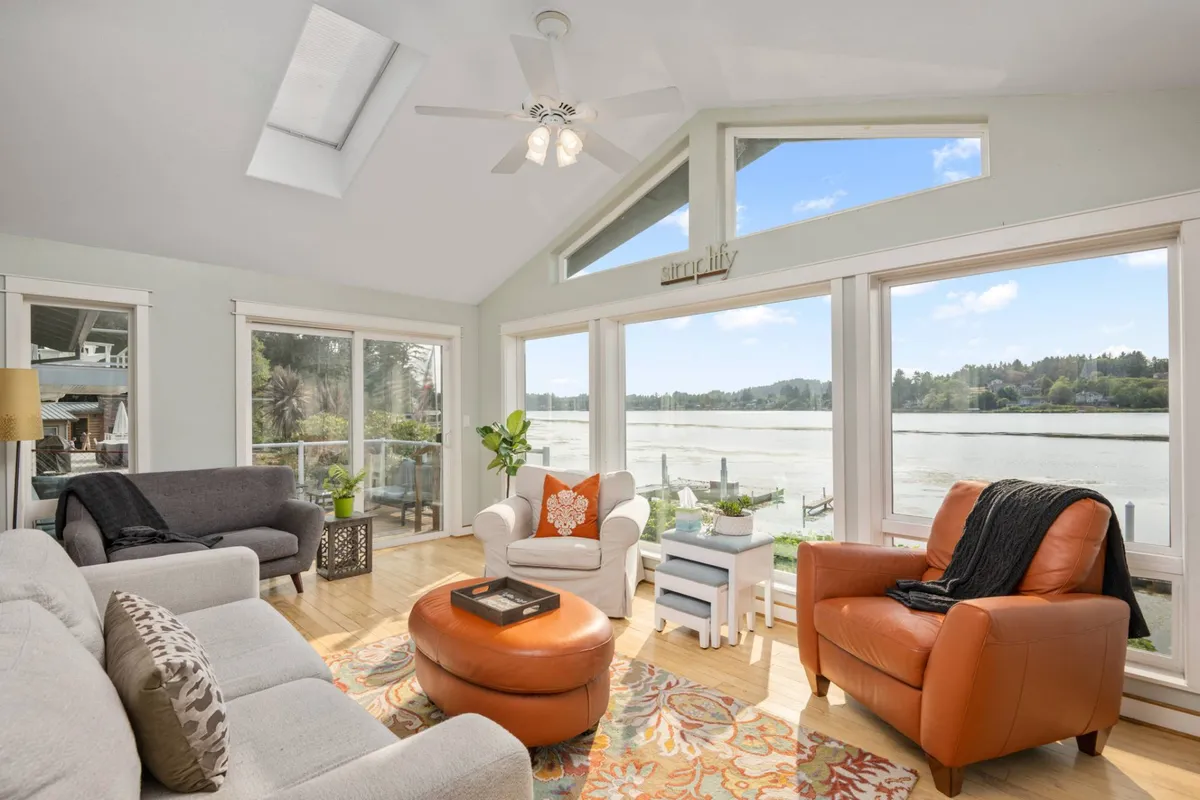 The image shows a bright, inviting living space with large windows framing serene lake views. Cozy chairs and a colorful area rug create a welcoming seating area, accented by decorative throw pillows. Natural light fills the room, enhancing the relaxing and homely atmosphere.